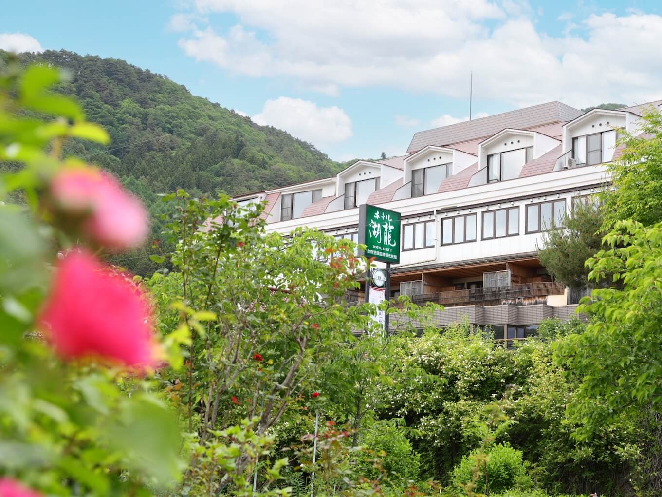 Hotel building with a green sign, set among lush greenery and flowering plants, hillside and blue sky in the background.