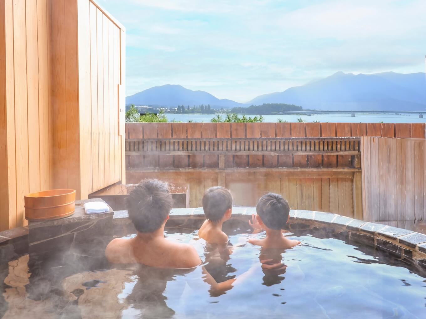 Three people soak in a steaming outdoor wooden hot tub on a balcony with a lake and mountains in the distance at dusk.