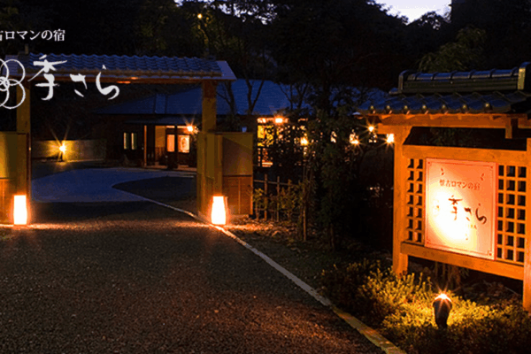 Night view of a traditional Japanese inn entrance (ryokan) with a wooden gate and warm lanterns illuminating the path. A lit signboard with Japanese text stands to the right.