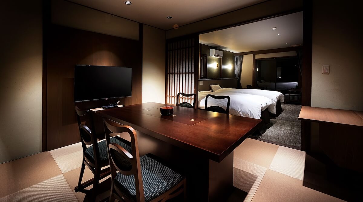 Hotel suite with a dark wooden dining table, four chairs, a wall-mounted TV, and two white beds in the background.|But context requires one sentence per alt text so separate entries are fine.
