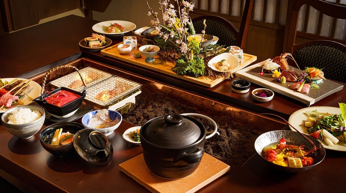 Elaborate Japanese multi-course meal on a dark wooden table, centered by a grill and a black pot, with sashimi, rice, and vegetables.