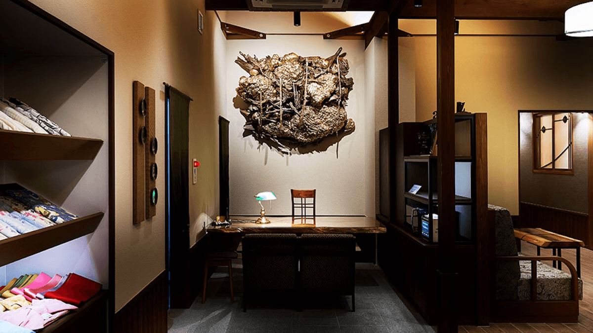 Indoor showroom workspace with a wooden desk and chair beneath a large textured wall sculpture, fabric swatches on the left.